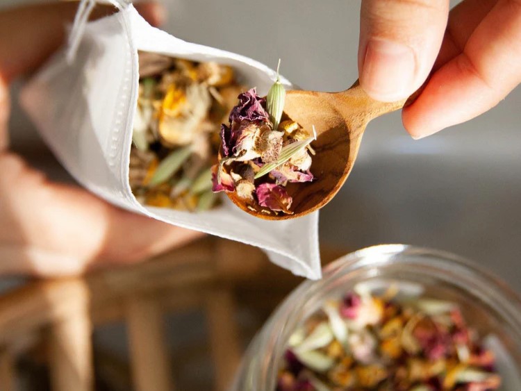 Hand holding a wooden spoon scooping loose herbal tea with rose petals from a paper pouch over a glass jar in warm light.